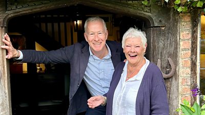 John Wilson and Dame Judi Dench stand in a doorway, smiling. A metal horseshoe is pinned to the wooden doorframe.