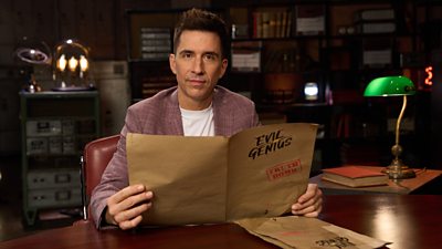 Photo of Russell Kane sat at a desk on the set of Evil Genius holding an Evil Genius portfolio marked "Truth Bomb". 