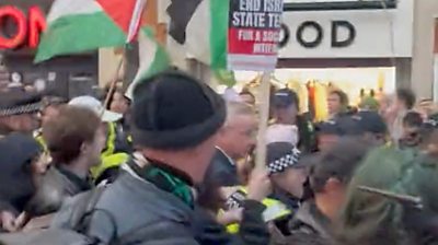 Michael Gove surrounded by police and protesters waving Palestinian flags