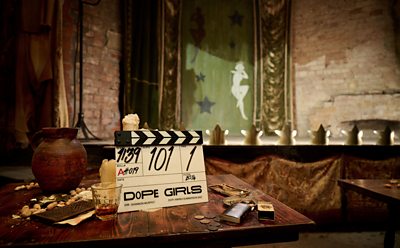 A Dope Girls clapperboard sits on a table in front of an illuminated theatre stage.