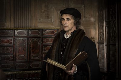 Mark Rylance as Thomas Cromwell in Wolf Hall series one. Stood in a grand, wood-panelled room, he holds a large, leather-bound book and is dressed in clothing from the Tudor period. 