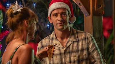 A scene from Death in Paradise 2023 Christmas episode featuring Doon Mackichan and Ralf Little having a conversation at a festive party - Ralph wears a red Santa hat. 