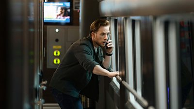 Joe Cole as Joe Roag - he stands in a train carriage, looking out the window whilst on his phone. He looks very concerned.