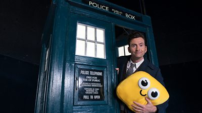 The Fourteenth Doctor (David Tennant) pops his head out the front door of the TARDIS, holding a yellow cushion with a smiley face on it.