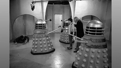 A black and white behind the scenes image from an early series of Doctor Who features filming of an episode featuring Daleks