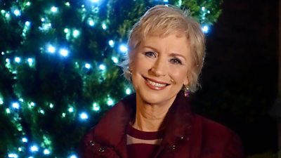 Sally Magnusson smiles to camera in front of a Christmas tree wrapped in fairy lights. 