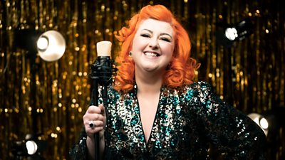 Michelle McManus smiles to camera, wearing a sequinned dress and holding a microphone. In the background are golden decorations and stage lights. 