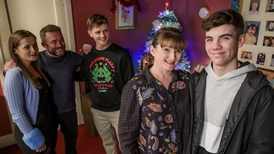 Photo from River City featuring five cast members wearing festive clothing. They all stand in a living room decorated for Christmas. 