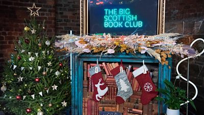 Photo of a fireplace adorned with festive decorations next to a Christmas tree. A screen above the fireplace mantel reads: “The Big Scottish Christmas Book Club”