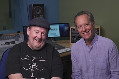 Johnny Vegas and Richard E Grant smile to camera from an audio recording studio. A computer in the background has ‘The Wombles’ written on screen. 