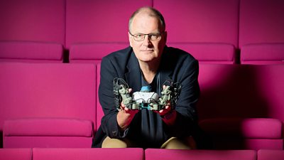 Mike Wooldridge, Professor of AI at Oxford, sits on bright pink seating holding a piece of technology in his hands