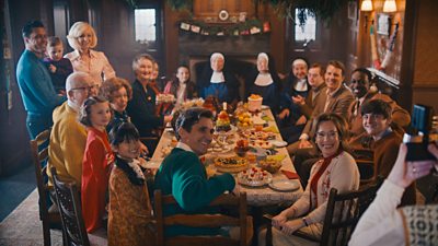 The cast of Call The Midwife sit around a dinner table piled with festive foods as someone takes a photograph