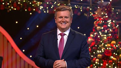 Aled Jones stands in front of a Christmas tree in a festive studio, smiling