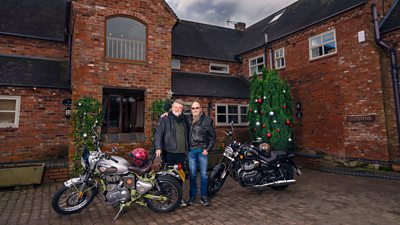 Si King and Dave Myers stand outside a large red brick house with their motorbikes