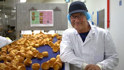 Gregg Wallace stands inside a factory beside a conveyor belt of Yorkshire puddings.