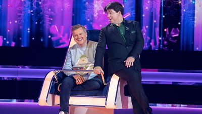 Michael McIntyre sits on the edge of Aled Jones' chair on The Wheel. Aled wears a Snowman jumper.