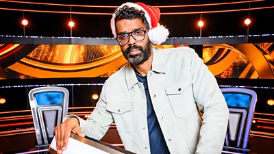 Romesh Ranganathan stands at the podium wearing a red Father Christmas hat