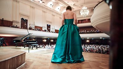 Aleksandra Świgut wears a long green gown as she stands on stage in a theatre, photographed from behind, facing the audience