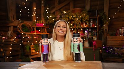 Stacey Solomon smiling to camera with two homemade nutcrackers in each hand. Background of Christmas decorations and fairy lights