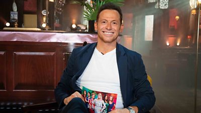 Joe Swash smiles to camera from the the set of the Queen Vic. He holds a card reading: “The Six Revealed” with photos of the six actors in the EastEnders storyline. 