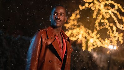 Nucti Gatwa as The Doctor, standing outside on a dark night in snow, with glittering lights in the background