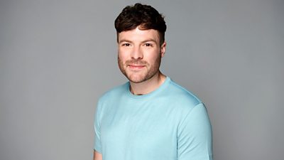Jordan North wears a blue t-shirt and smiles. He's pictured against a grey background.