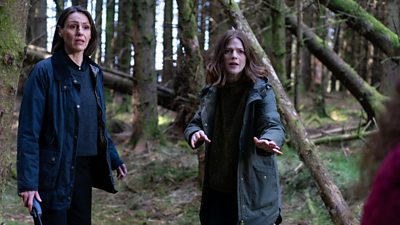 Amy Silva (Suranne Jones) and Kirsten Longacre (Rose Leslie) stand in a forest looking concerned. The pair listen to character off-camera. 