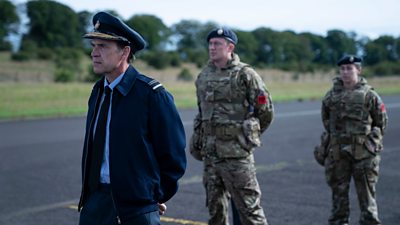 Marcus Grainger (Dougray Scott) wearing a military cap and jacket over a shirt and trousers. Stood on a runway in front of two other characters in military uniform. 