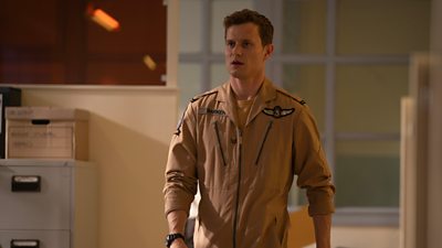 Callum Barker (Chris Jenks) looks shocked as he walks into an office, wearing air force overalls. 