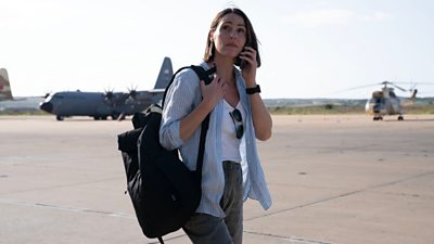 Amy Silva (Suranne Jones) walks on a runway carrying a backpack and holding a phone to her ear. There are large military planes in the background. 