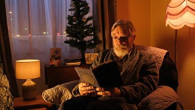 Image of James Fleet appearing in the Beyond Paradise Christmas special 2023. He reads a book in an armchair, with a small Christmas tree and fairy lights decorating the window behind him. 