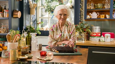 Mary Berry stands in a festively decorated kitchen, resting her arms on a kitchen island and smiling. In front of her on the counter is a Buche de Noel, a French Christmas cake made from a thin layer of sponge cake that is rolled up and decorated to resemble a Yule log