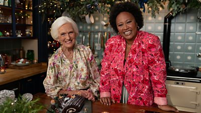 Mary Berry and Emeli Sandé stand in the kitchen, resting their hands on the kitchen island. In front of them on the counter is Mary's Buche de Noel, a French Christmas cake made from a thin layer of sponge cake that is rolled up and decorated to resemble a Yule log