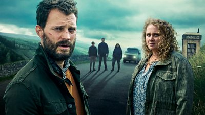 Key art for The Tourist series two featuring Jamie Dornan and Danielle Macdonald looking to camera as they stand on a remote country road. Three unidentifiable figures are stood in the background next to a truck. 