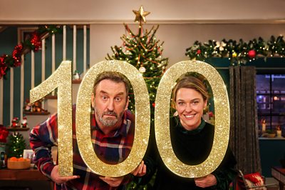 Lee Mack and Sally Bretton holding a gold 100 with Christmas decorations in the background