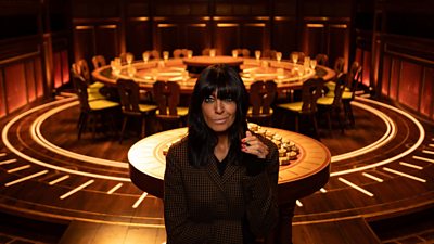 Claudia stands in front of a small round table piled with gold discs and a large round table surrounded by chairs