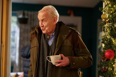 Geoffrey (Geoffrey Whitehead) holds a cup as he chats to someone off-screen. 