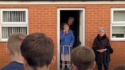 A woman standing in the doorway of her house as people sing carols outside