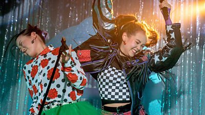 Two girls dance on a stage holding microphones