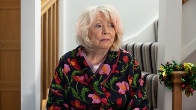 Alison Steadman sits on the end of the stairs in a floral dressing gown.