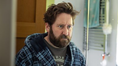 Jim Howick looks at himself in a bathroom mirror. He wears a black t-shirt beneath a blue patterned dressing gown.