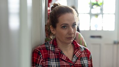 Katherine Parkinson leans against a white door in red plaid patterned pyjamas.