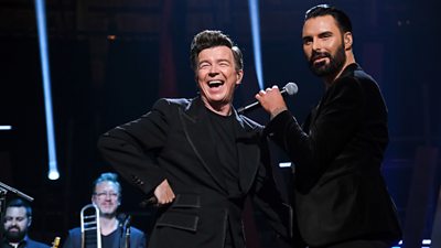 Rick Astley and Rylan stand on stage together. Rick is laughing and Rylan looks towards the crowd.