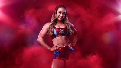 Dynamite stood against a red background, with both hands on their hips 