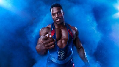 Nitro stood against a blue background, pointing to the camera 