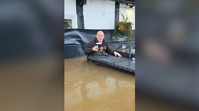 A man sitting in water that is up to his hips holding a pint of beer