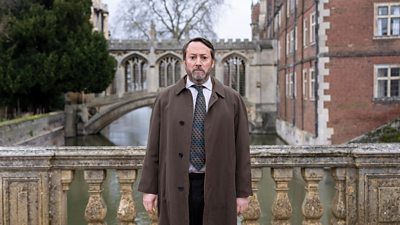 David Mitchell as Ludwig. He stands on a stone bridge over a river, wearing a long brown coat, white shirt and tie, looking serious.
