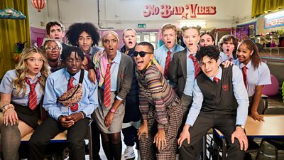 Group photo from Bad Education series five, featuring students in uniform stood alongside teachers. A poster in the background reads: “No bad vibes” 
