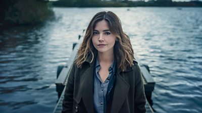Jenna Coleman as character Ember Manning. She looks to the camera standing on a wooden pier with a lake in the background