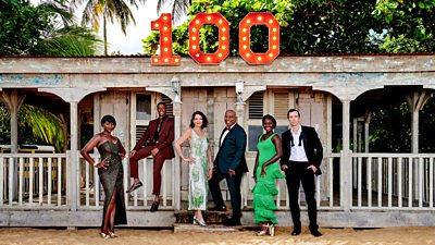 Ginny Holder, Tahj Miles, Elizabeth Bourgine, Shantol Jackson and Ralf Little on the terrace of a wooden building on a sand-covered beach. A large “100” made of lightbulbs is attached to the front of the building. 